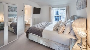 Une chambre avec un grand lit, un téléviseur fixé au mur, une fenêtre avec des rideaux et une porte donnant sur une autre pièce.
