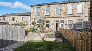 Une maison en pierre à deux étages, avec un petit jardin devant, une clôture en bois et un chemin pavé menant à l’entrée.