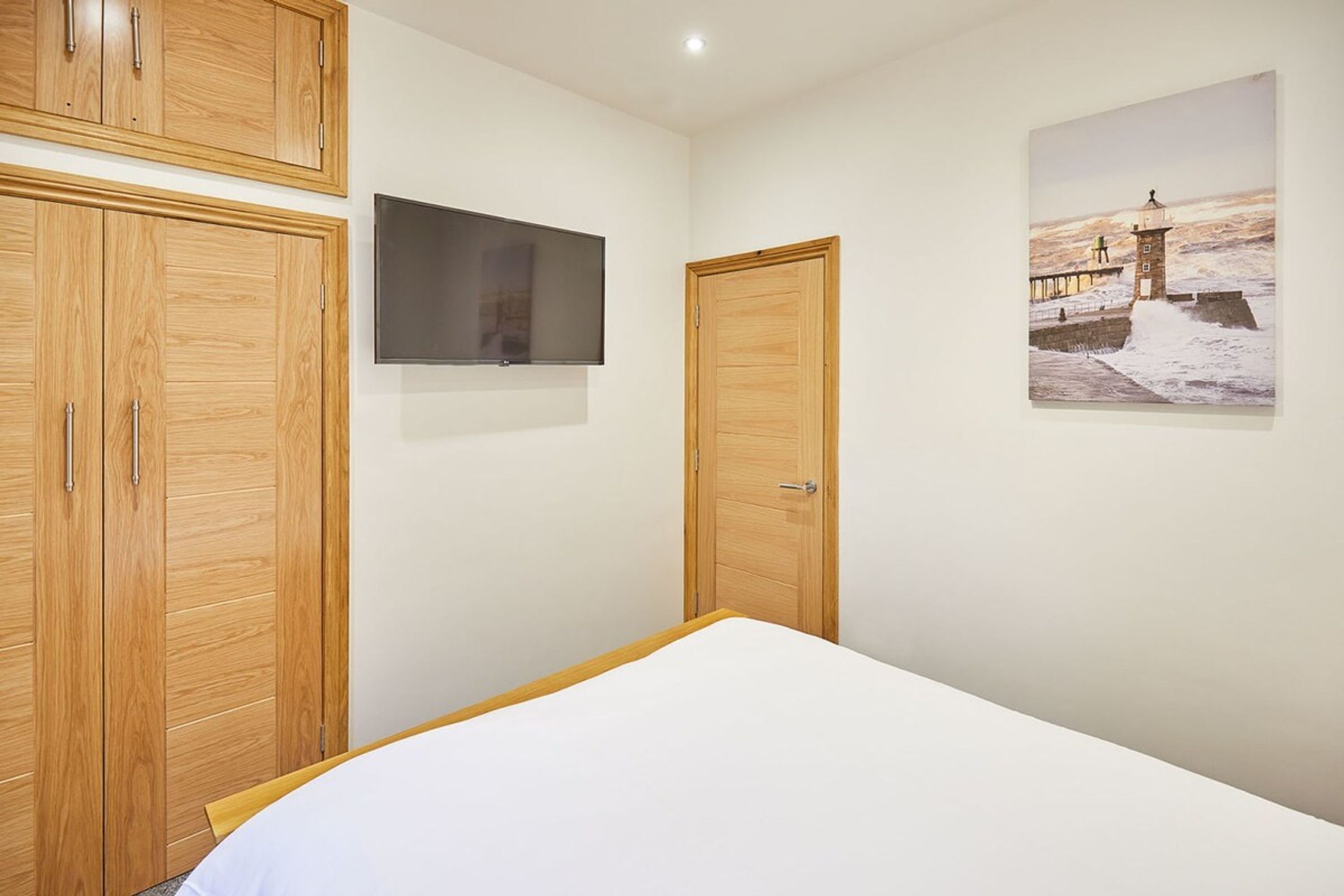 A bedroom with a bed, wooden wardrobe, a television, and a painting of a coastal scene.