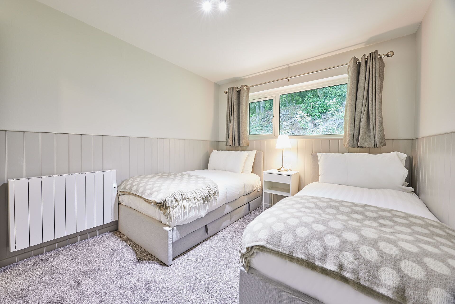 A bedroom with two beds, a window with curtains, a bedside table with a lamp, and a radiator.
