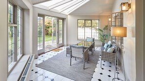 Ein sonnendurchflutetes Zimmer mit einem großen Fenster, einem Korbsessel, einem kleinen Tisch und einer Stehlampe.