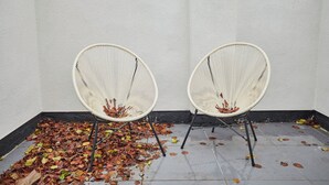 Two white wicker chairs with metal legs, placed against a white wall with a pile of fallen leaves on the floor.