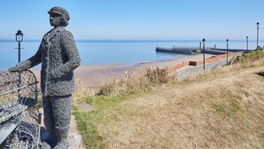Eine Statue einer Person, die am Meer steht, mit einem Steg im Hintergrund.