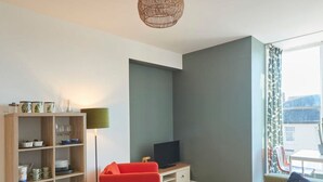 Ein modernes Wohnzimmer mit Holzboden, einem grünen Sofa, einem roten Sessel, einem Fernseher und einem Bücherregal.