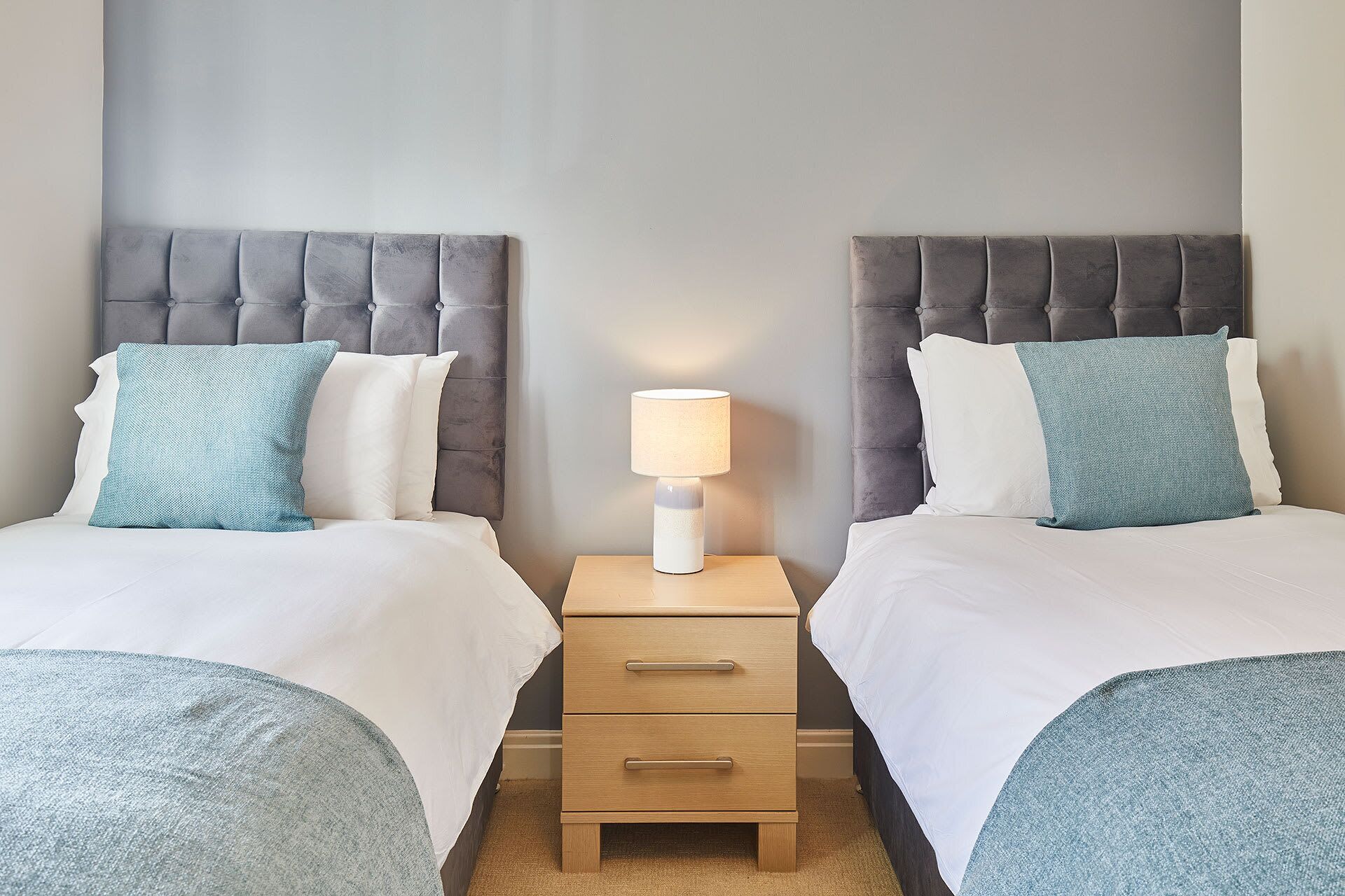 A hotel room with two beds, each with a grey upholstered headboard, a bedside table with a lamp, and a small wooden drawer unit.