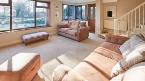 A spacious living room with large windows, a brown leather sofa, a matching ottoman, and a staircase leading to an upper floor. - Host Stay The Chimneys (Alnwick)