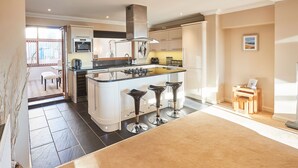 A modern kitchen with a central island, bar stools, and a dining area with a table and chairs. - Host Stay The Chimneys (Alnwick)