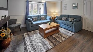 A living room with two blue sofas, a wooden coffee table, a grey armchair, a fireplace, and a large window with blue curtains.