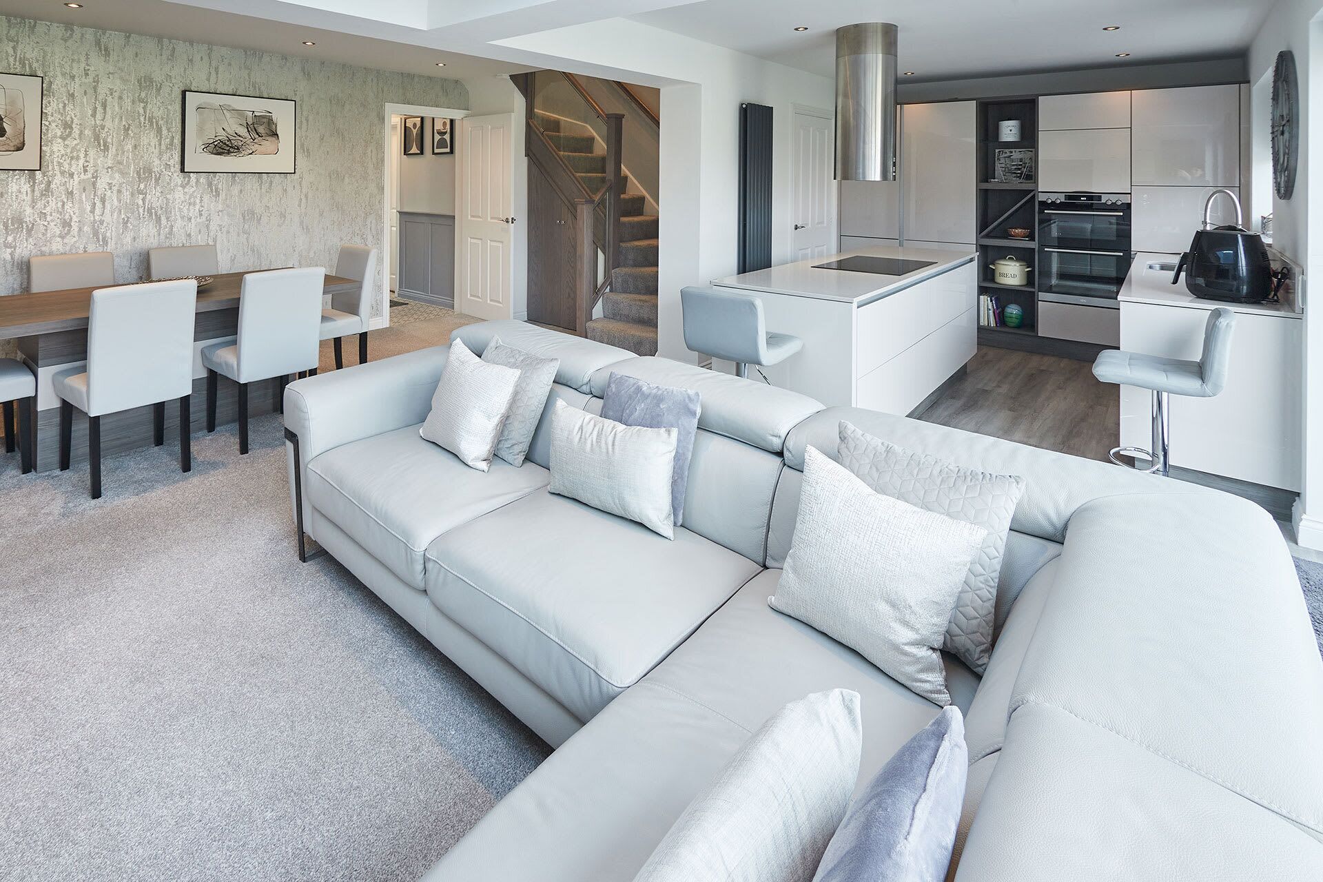 A modern living room with a light-colored sectional sofa, a dining area with white chairs, and a kitchen with stainless steel appliances.