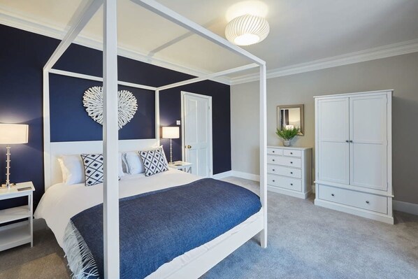 A four-poster bed with a blue bedspread, a white wardrobe, a nightstand with a lamp, and a decorative heart on the wall.