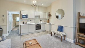 A modern kitchen with white cabinets, a built-in oven, and a gas stove. There is a bench with a striped cushion and a round mirror on the wall. - Host Stay Silver Beach (Whitby)
