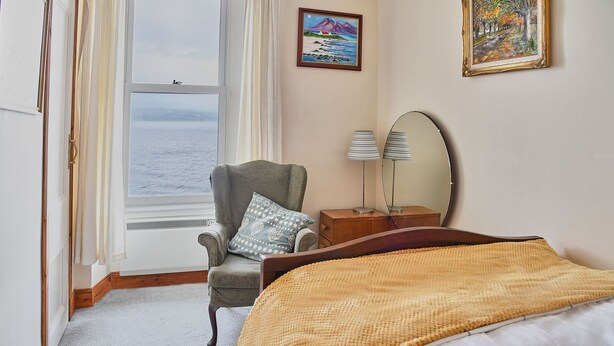 Une chambre avec une grande fenêtre, une chaise, une table de chevet avec une lampe et un lit recouvert d’une couverture jaune.