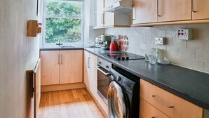 Une cuisine avec des meubles en bois, un plan de travail noir, une fenêtre donnant sur un espace verdoyant, un lave-linge et une bouilloire.