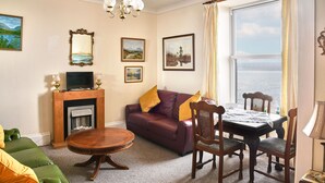 Un salon avec une cheminée, un canapé, une table à manger avec des chaises et une vue sur la mer, visible à travers une grande fenêtre.