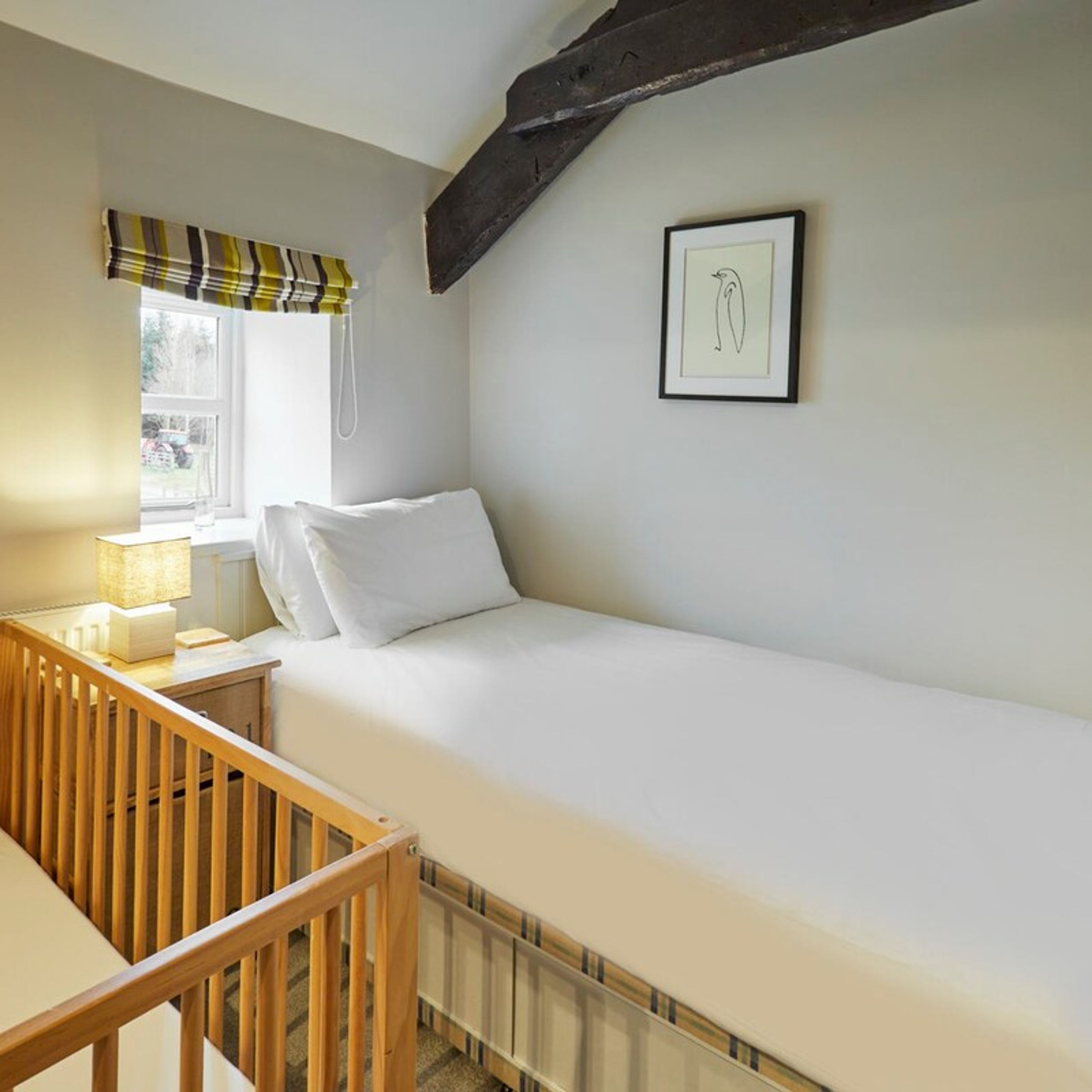 A single-bed room with a wooden crib, a bedside table with a lamp, a window with a striped curtain, and a framed picture on the wall.