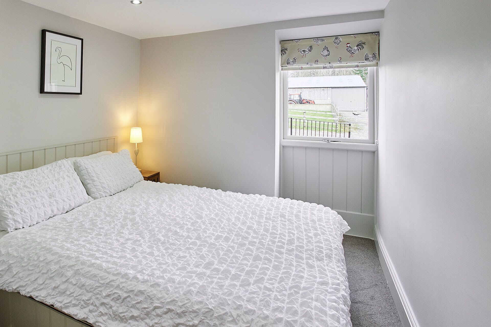 A bedroom with a large bed, a bedside lamp, a framed picture on the wall, and a window with a view of a farm.
