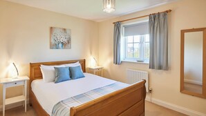 A bedroom with a wooden bed, white nightstands, a bedside table, a window with curtains, a radiator, and a painting on the wall. - Host Stay Driftwood (Chathill)
