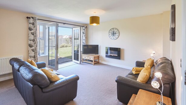 A living room with two black leather sofas, a wooden coffee table, a flat-screen TV, a clock, and a sliding glass door leading to a garden. - Host Stay Driftwood (Chathill)