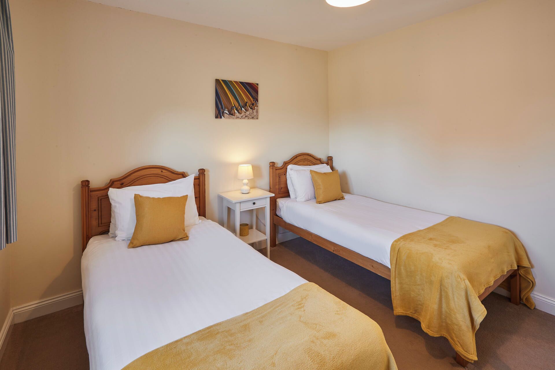Habitació d'hotel amb dos llits individuals, cadascun amb un capçal de fusta, roba de llit blanca i una manta groga. Hi ha una petita taula de nit amb una làmpada i un quadre sobre el llit esquerre.