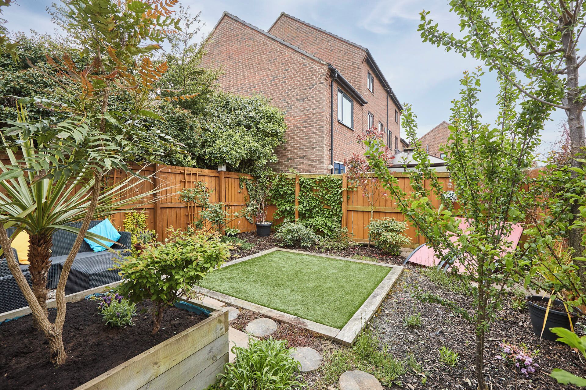 A well-maintained backyard with a lawn, various plants, a wooden fence, and a brick house in the background.
