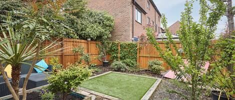 A well-maintained backyard with a lawn, various plants, a wooden fence, and a brick house in the background.