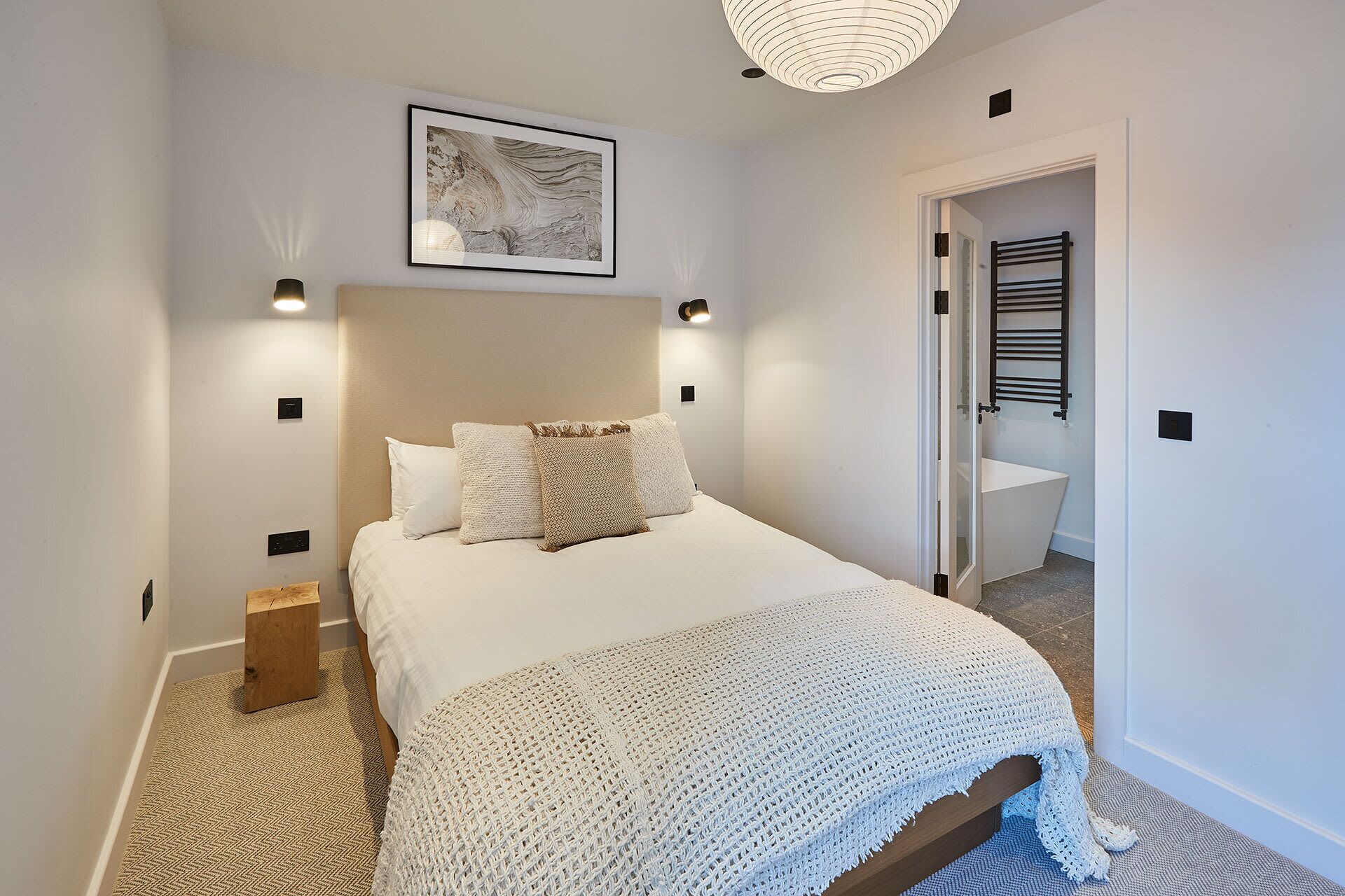 Un dormitorio con una cama grande, una obra de arte en la pared, una mesita de noche y una puerta que da al baño.