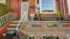 A red brick house with a white door, a bay window, and a small garden with various plants and flowers. - Host Stay High Street (Saltburn-by-the-Sea)