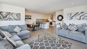 Ein modernes Wohnzimmer mit zwei grauen Sofas, einem Glastisch und einem gemusterten Teppich. Im Hintergrund ist eine Küche mit weißen Schränken und ein Essbereich mit Tisch und Stühlen zu sehen.