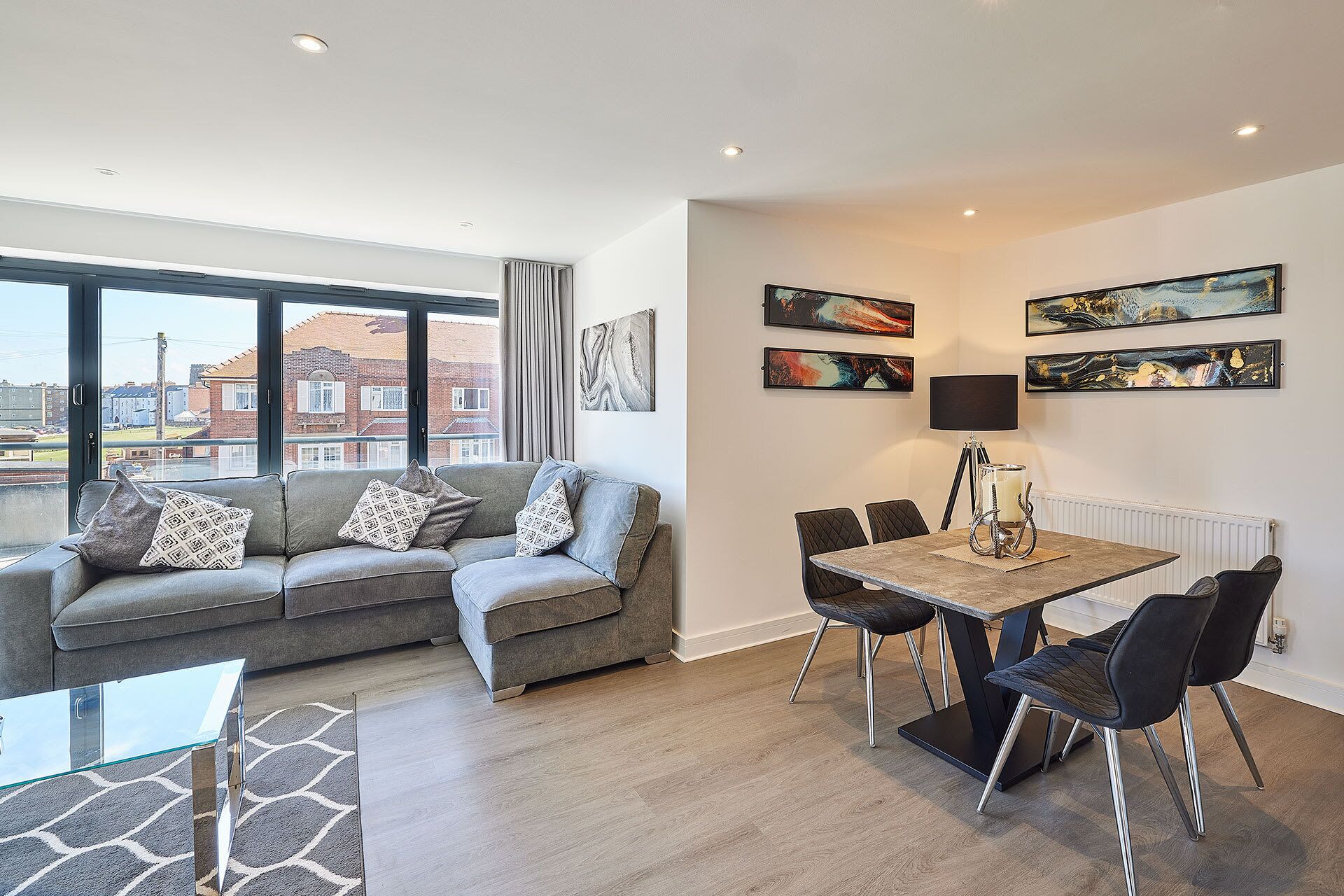 A modern living room with a grey sofa, a glass coffee table, and a dining area with a wooden table and black chairs. There are abstract paintings on the wall and a large window with a view of buildings.