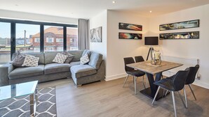 Ein modernes Wohnzimmer mit grauem Sofa, einem Glastisch und einem Essbereich mit Holztisch und schwarzen Stühlen. An den Wänden befinden sich abstrakte Gemälde und ein großes Fenster mit Blick auf Gebäude.