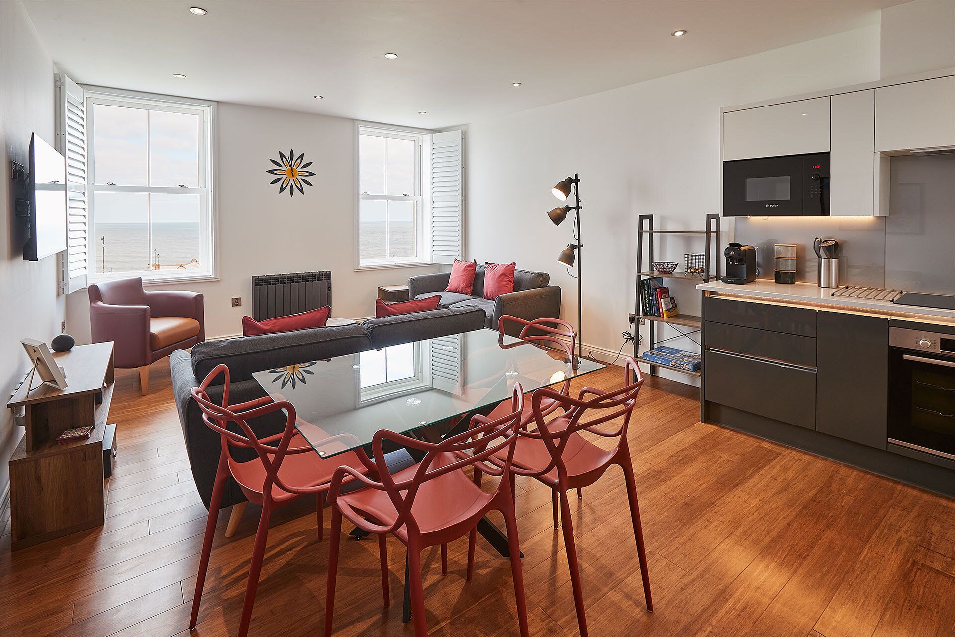 Espaço moderno com mesa de jantar de vidro, cadeiras vermelhas, sofá cinza e máquina de café preta.
