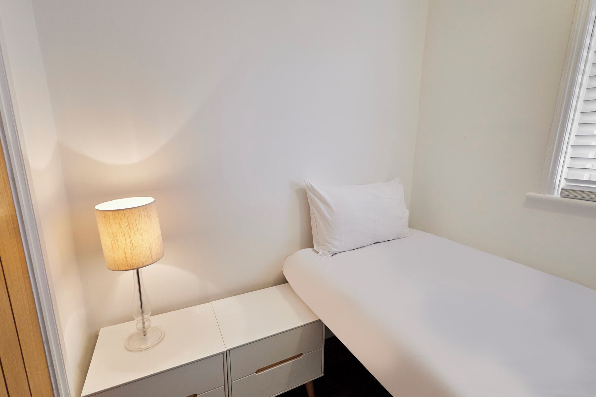 Un dormitorio pequeño y ordenado con una cama individual, una mesita de noche con una lámpara y una ventana con persianas.
