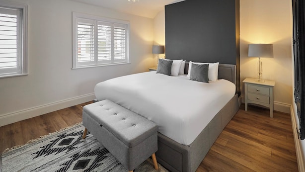 Un dormitorio moderno con una cama grande, un banco gris, una araña y una mesita de noche con lámpara.