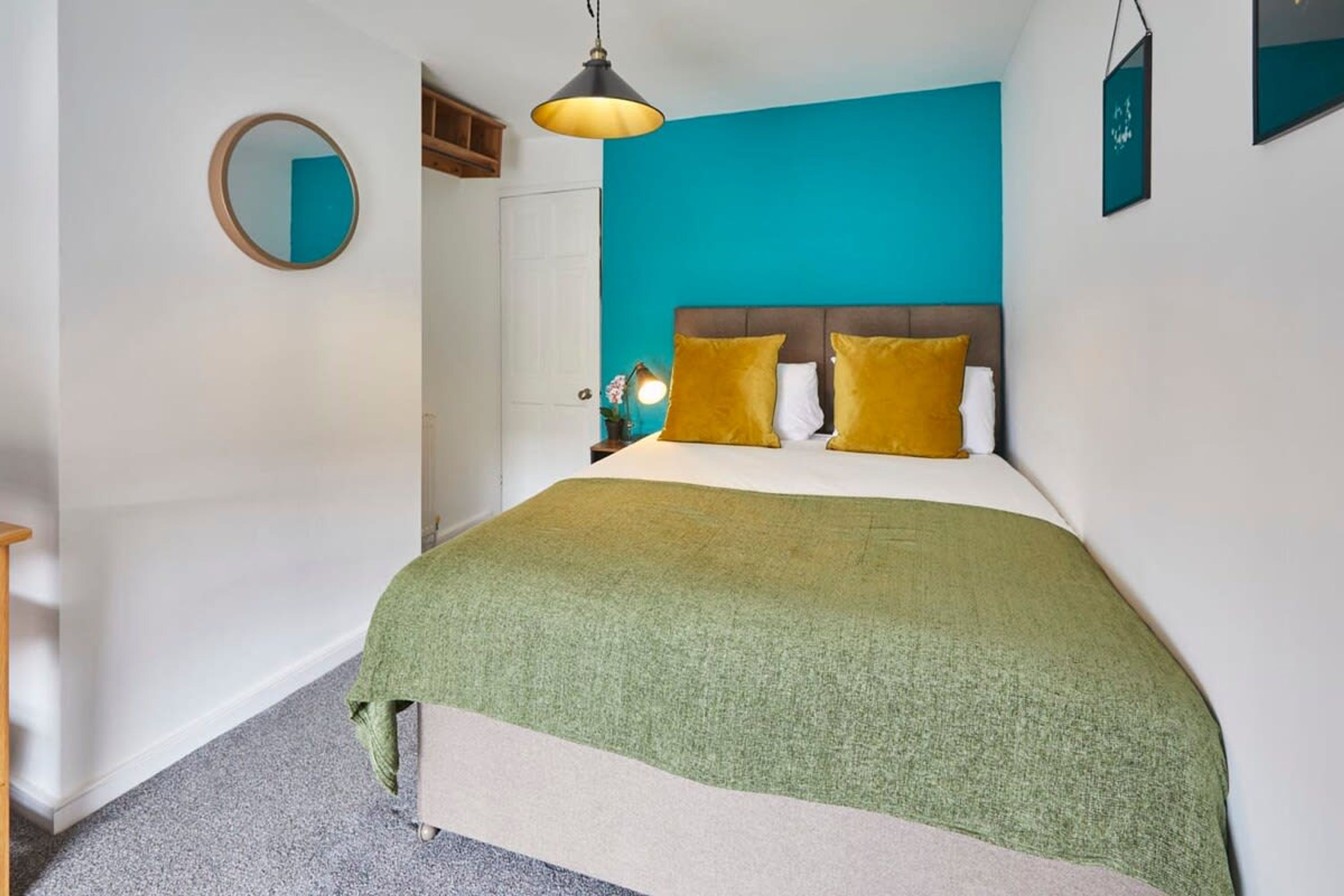 A bedroom with a teal accent wall, a round mirror, a bed with green and white bedding, and a pendant light.