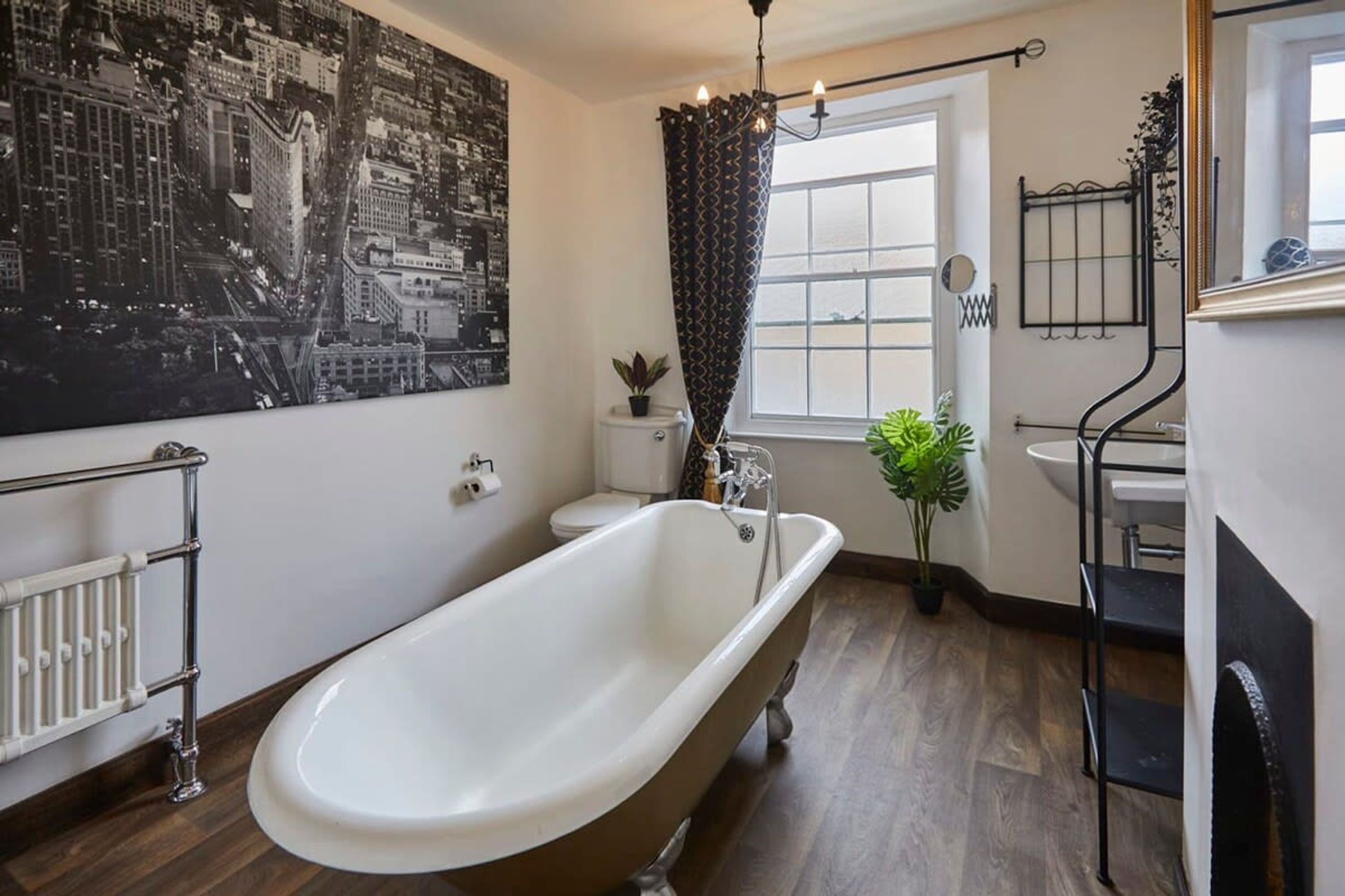 A bathroom with a freestanding bathtub, a radiator, a toilet, a window with curtains, and a wall-mounted mirror.