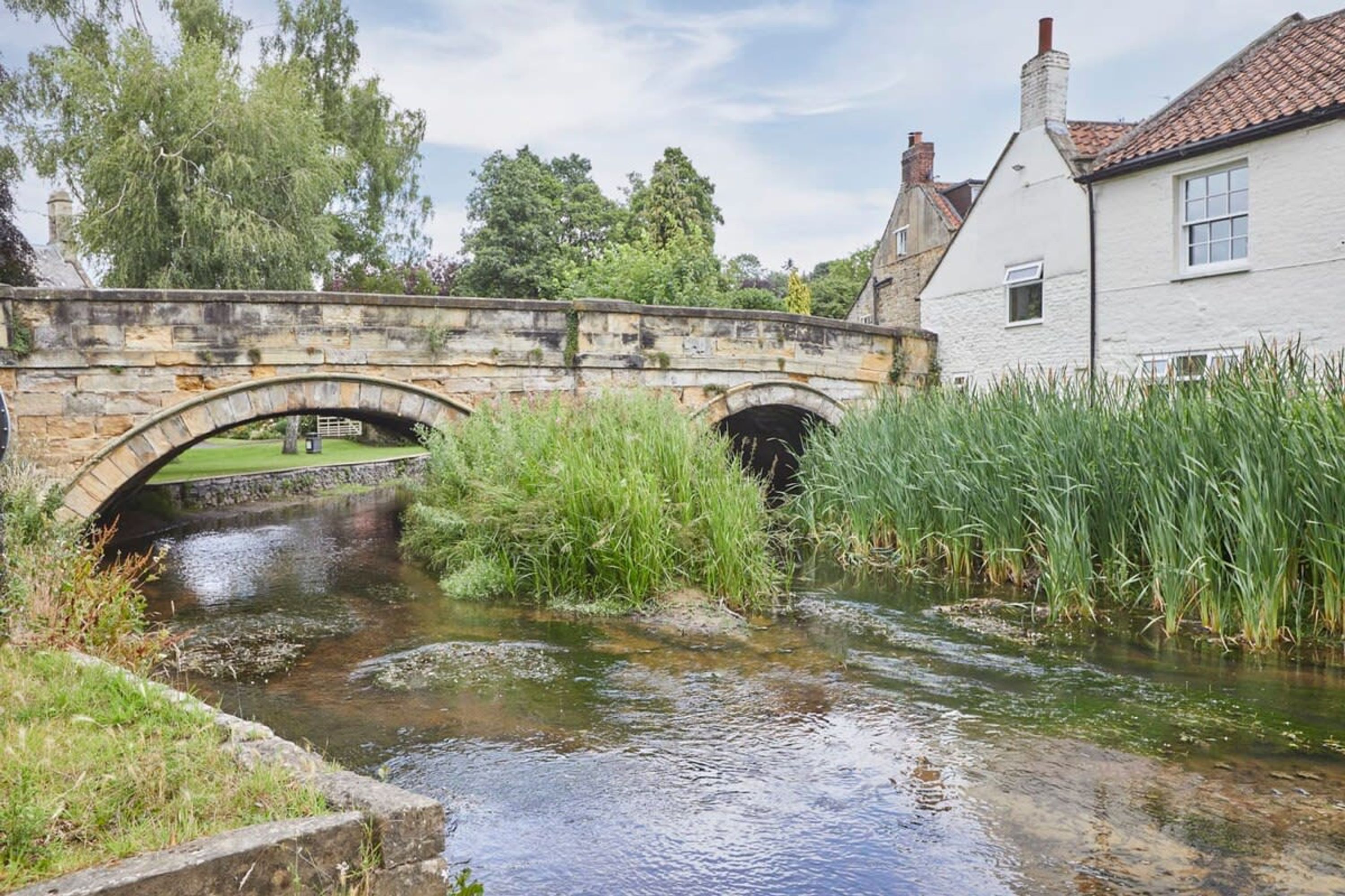 A stone bridge over a narrow stream with grass on either side, a white building with a red-tiled roof in the background.