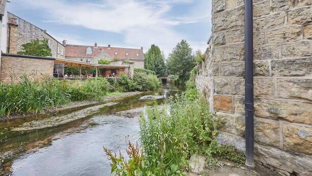 A narrow water channel with stone buildings on either side, a stone wall on the right, and a building with a red roof in the background. - Host Stay Beckside (Pickering)