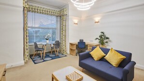 A living room with a blue sofa, a coffee table, a dining table with chairs, a desk, and a potted plant. - Host Stay Thamesview (Staines)