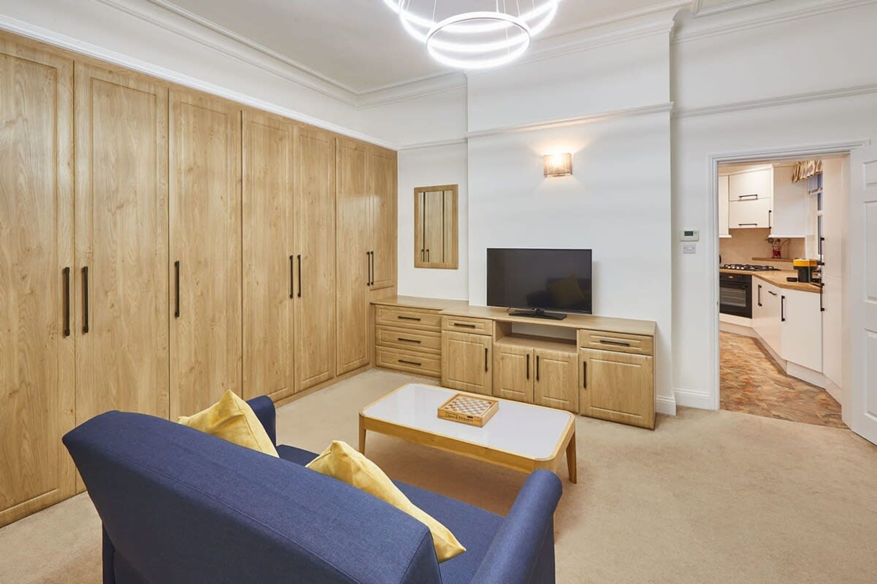 A living room with a sofa, coffee table, TV, wooden cabinets, and a kitchen in the background.