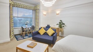 A modern hotel room with a bed, a blue sofa with yellow cushions, a small desk, a chair, a table with a chessboard, and a large window with blinds. - Host Stay Thamesview (Staines)