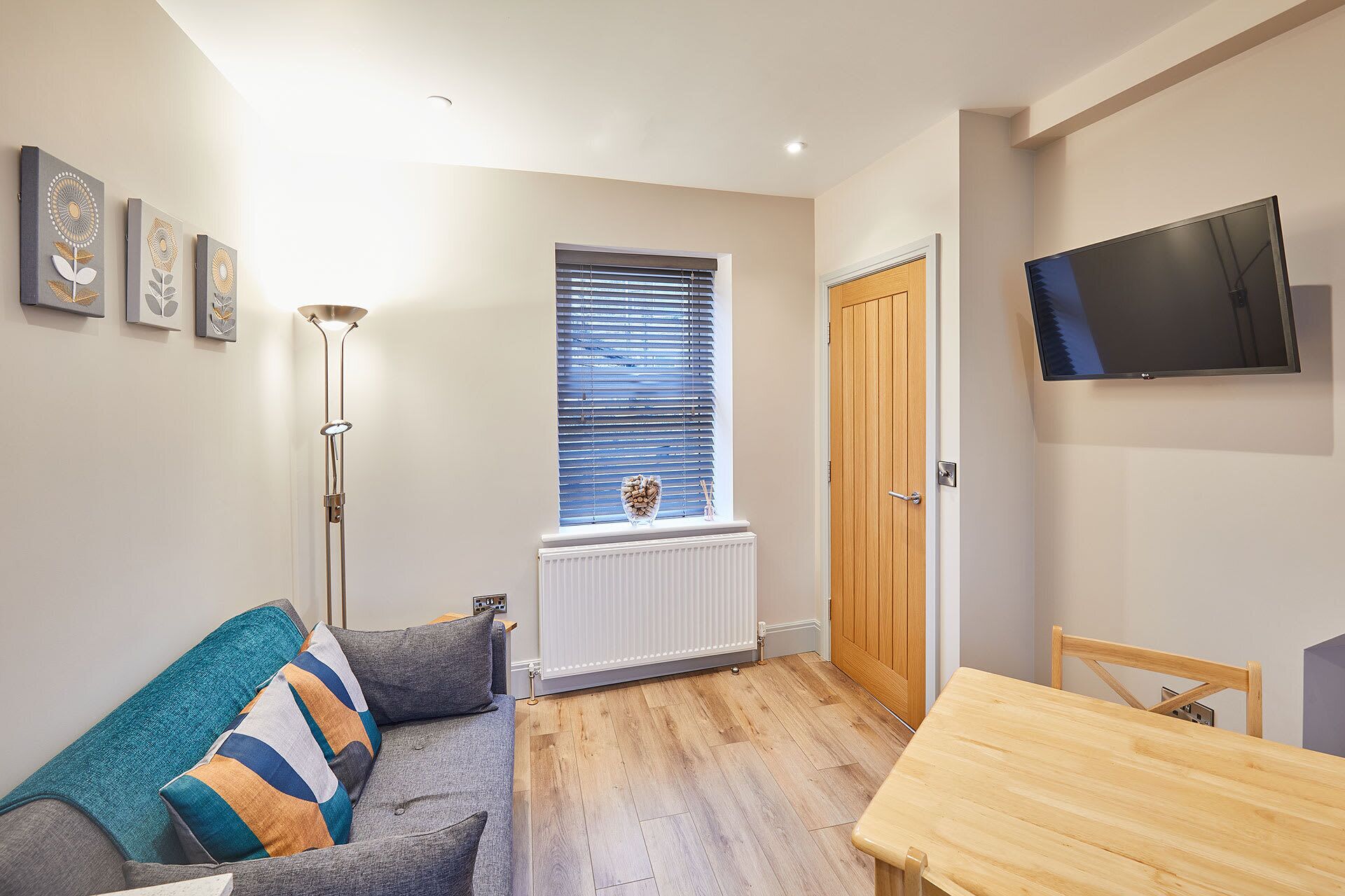 A modern living room with a grey sofa, a wooden dining table, and a TV mounted on the wall.