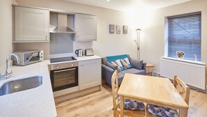 Eine kleine Küche mit Mikrowelle, Toaster, Wasserkocher und Backofen. Ein Essbereich mit Tisch und Stühlen, eine Couch und ein Fenster mit Jalousien.