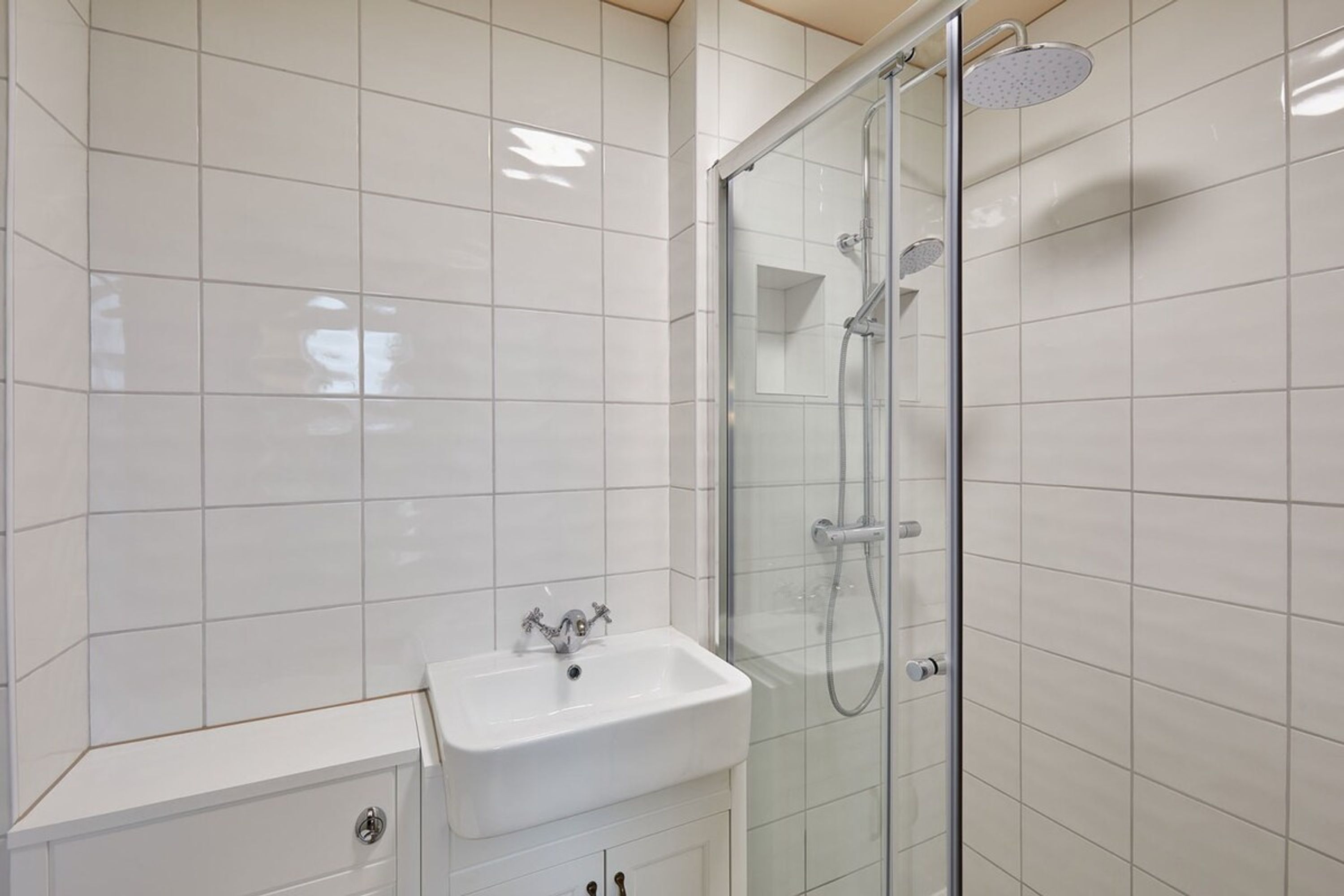 A modern bathroom with white tiles, a glass-enclosed shower, and a white sink with a cabinet below.
