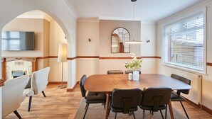 Un comedor con mesa de madera, seis sillas, un florero con flores, chimenea, televisor y un amplio ventanal con persianas.