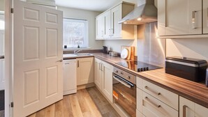 Une cuisine moderne avec des meubles blancs, un plan de travail en bois, un four encastré et une fenêtre équipée de stores.