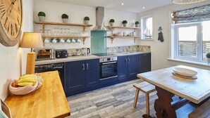 A modern kitchen with dark blue cabinets, a wooden dining table, and a large clock on the wall. - Host Stay Mumbles Cottage (Whitby)