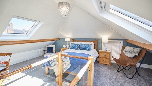 A bedroom with a skylight, a bed with blue bedding, a wooden chair, a nightstand, and a lamp. - Host Stay Mumbles Cottage (Whitby)
