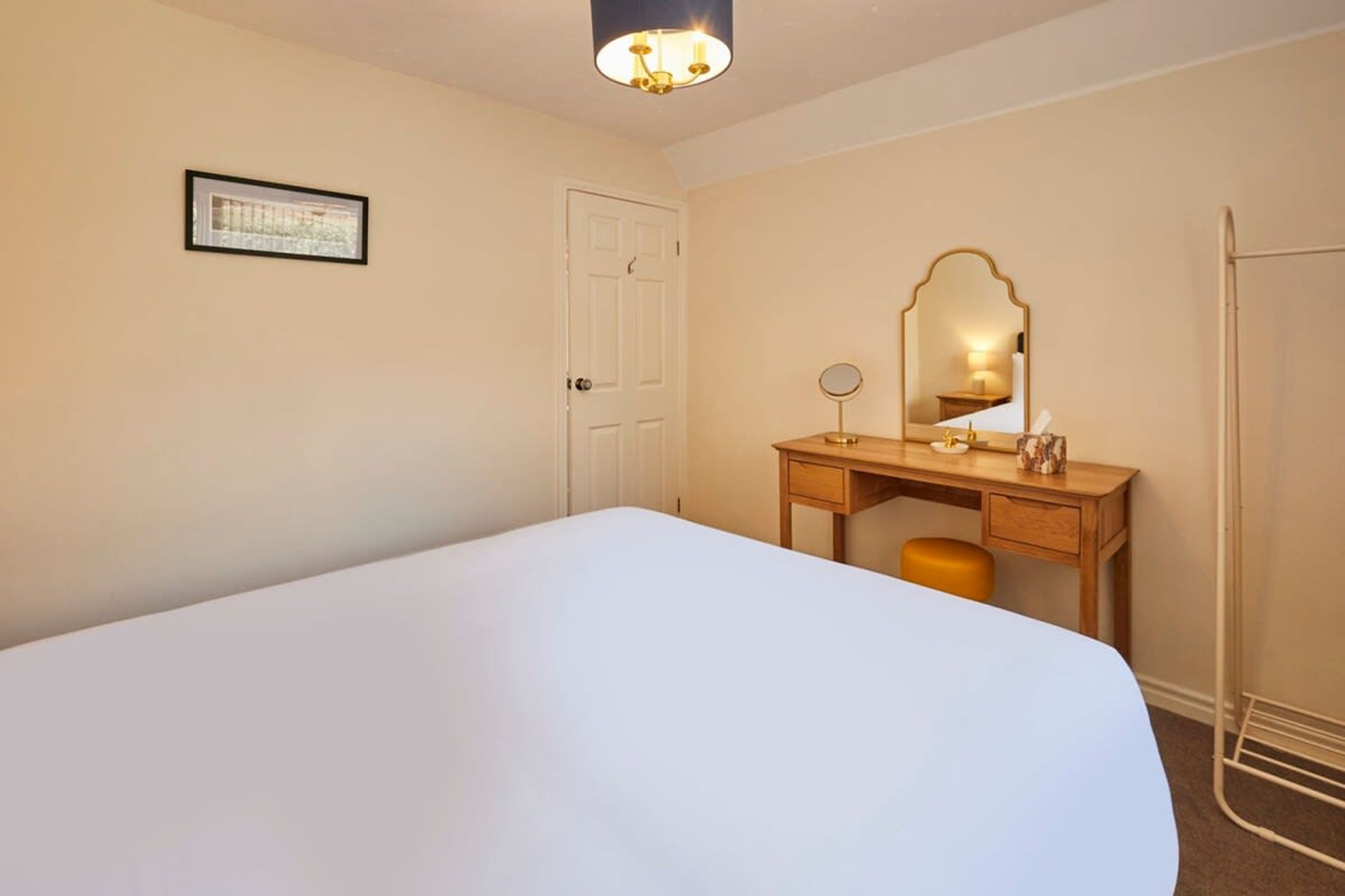 A hotel room with a bed, a wooden desk with a mirror, a small stool, and a door.
