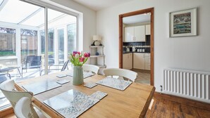 A dining area with a wooden table, white chairs, and a vase with flowers. There is a kitchen in the background with cabinets and a radiator. - Host Stay Cleveland Rest (Saltburn-by-the-Sea)
