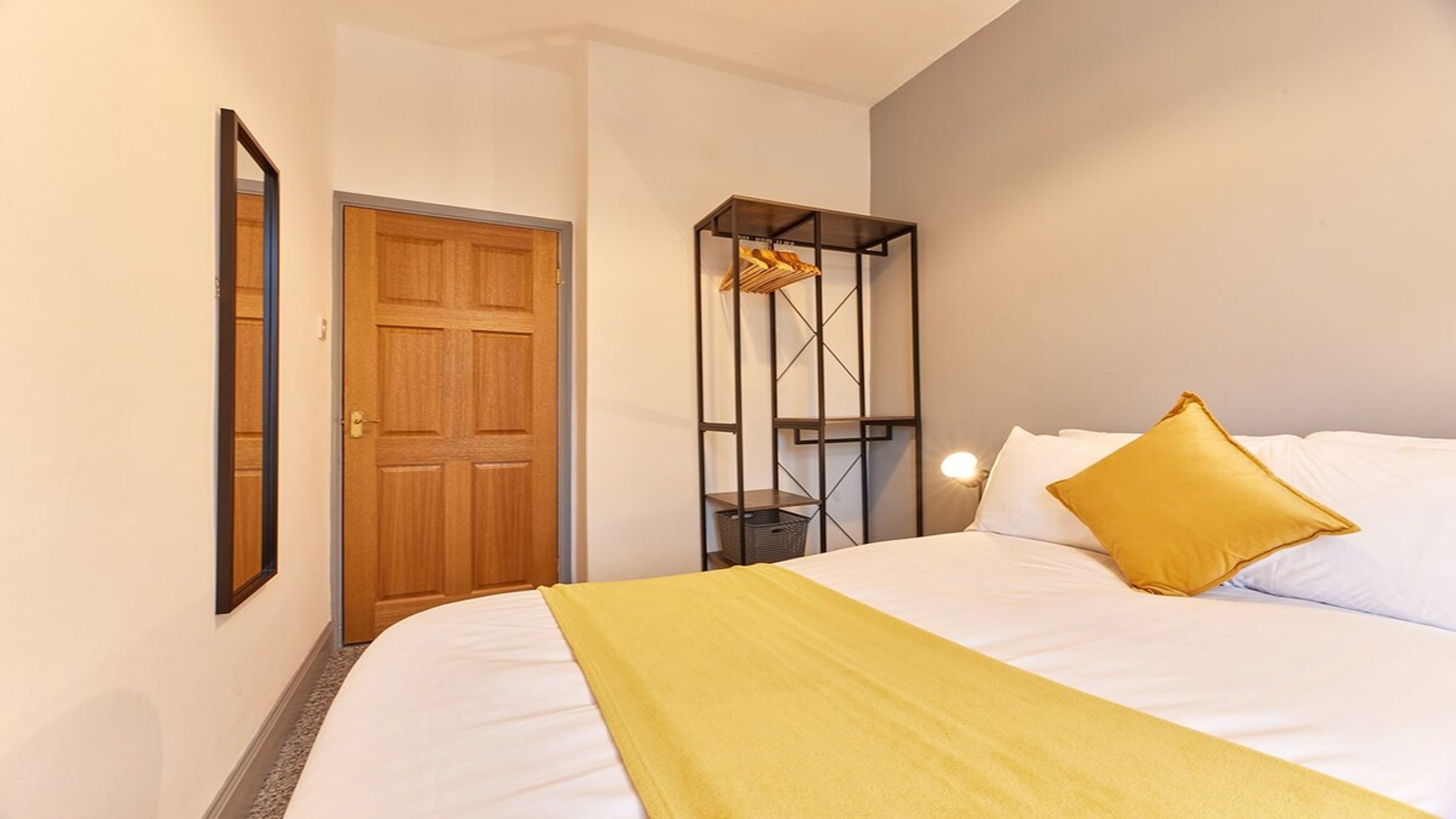 A hotel room with a bed, a wooden door, a wardrobe with hanging clothes, and a nightstand with a lamp.